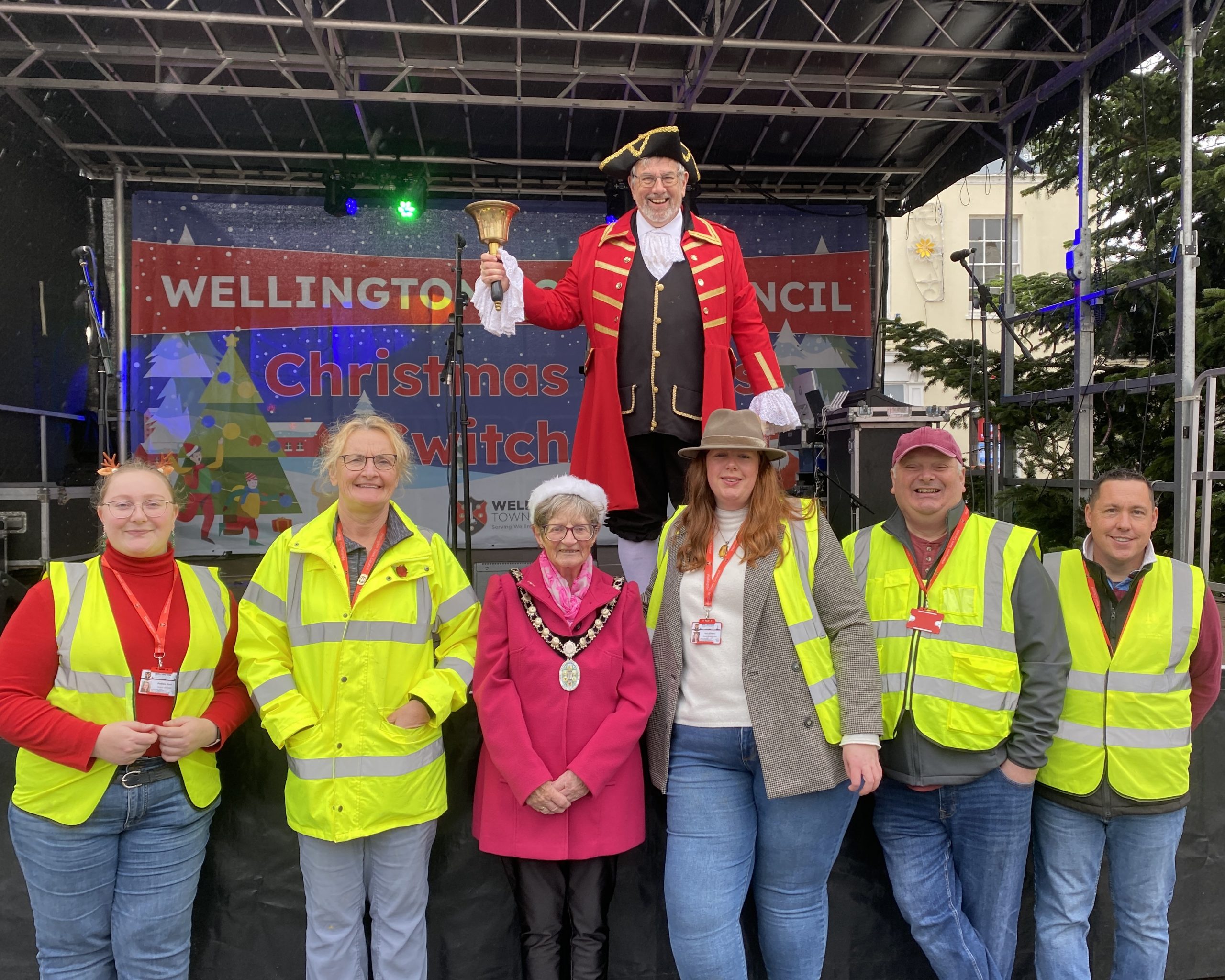 The Mayor of Wellington poses with five members of staff from Wellington Town Council, the staff members wear hi-vis. Town Crier Andrew Norris stands behind the group and poses onstage.