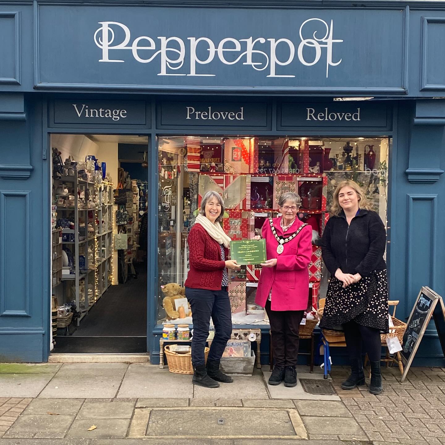 Three people stand in front of a shop window with a certificate.