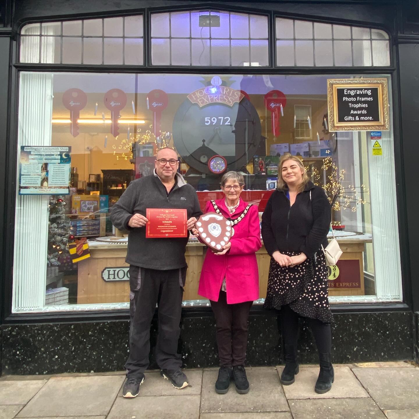 Three people stand in front of a shop window with a certificate and an award shield.