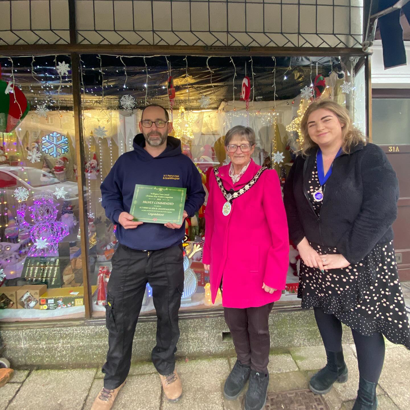 Three people stand in front of a shop window with a certificate.